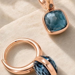 Pode incluir: Um par de brincos de argola em ouro rosa, cada um com uma pedra preciosa azul-escura facetada de corte quadrado, engastada numa armação de metal polido. Os brincos possuem um mecanismo de fecho de dobradiça e um tom azul profundo e translúcido.