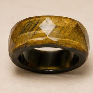 May include: A wide band ring crafted from golden-brown tiger's eye gemstone. The exterior surface features a faceted, geometric cut with a polished finish, showcasing the natural chatoyant bands and dark inclusions characteristic of the stone.