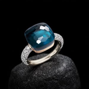 May include: A square-cut, teal gemstone ring with a gold band and diamond accents. The ring's setting is gold, and the band is silver. The ring is displayed on a dark, textured surface against a black background.
