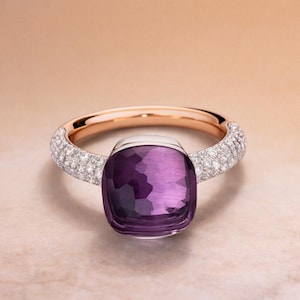 Può includere: Un anello in oro rosa con un grande e sfaccettato centro di pietra preziosa viola. La fascia è incastonata con piccole gemme trasparenti. L'anello è posto su uno sfondo morbido e neutro, evidenziando l'eleganza e il design del gioiello.
