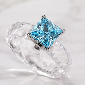 Puede incluir: Un anillo con una banda transparente facetada de aspecto cristalino y texturizado. Una piedra preciosa azul brillante de corte cuadrado está engastada en una montura de metal plateado en la parte superior, creando un contraste entre los elementos.
