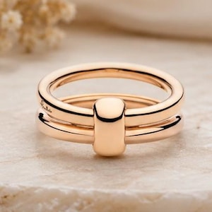 May include: A rose gold ring featuring two parallel bands connected by a single, smooth, vertical cylindrical element. The metal has a polished, reflective finish, and a small, delicate script engraving is visible on the side of the upper band.