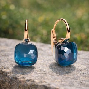 Può includere: Un paio di orecchini color oro con pietre preziose blu scuro, quadrate e sfaccettate. Gli orecchini hanno una chiusura a leva e sono esposti su una superficie di pietra contro uno sfondo verde sfocato. Le gemme riflettono la luce.