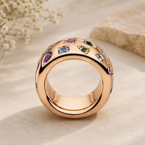 Könnte beinhalten: Ein breiter, polierter Ring aus Roségold mit einer Vielzahl von flächenbündig gefassten, mehrfarbigen Edelsteinen in verschiedenen Formen, darunter Rund-, Tropfen- und Marquise-Schliffe in Blau-, Grün-, Lila- und Rosatönen.
