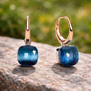 Op de afbeelding: Een paar elegante oorbellen met een vierkante, facetgeslepen, blauwgroene edelsteen hanger. De oorbellen hebben een goudkleurige oorring en een zilverkleurige basis. De oorbellen worden getoond op een natuurstenen oppervlak met een vage groene achtergrond.