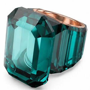 Könnte beinhalten: Ein Ring mit einem großen, smaragdgeschliffenen blaugrünen Kristall, der in ein roségoldenes Metallband gefasst ist. Der Kristall hat ausgeprägte Facetten, die das Licht reflektieren und tiefe blaugrüne Töne im Stein erzeugen.