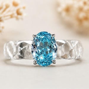 Puede incluir: Un anillo con una piedra preciosa azul brillante de corte ovalado sujeta por cuatro garras plateadas. La banda está hecha de un material transparente y facetado que crea una apariencia texturizada de múltiples caras.