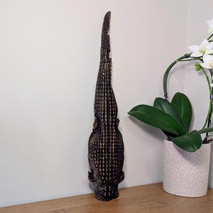 May include: A tall, dark brown alligator sculpture with a textured surface. The sculpture is made of a dark brown material and is placed on a wooden surface. A white orchid plant in a pink textured pot is in the background.