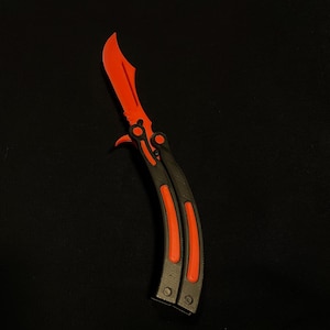 Könnte beinhalten: Ein Butterflymesser mit leuchtend roter Klinge und Akzenten. Der Griff ist schwarz mit roten Streifen. Das Messer wird vor einem schwarzen Hintergrund gezeigt.