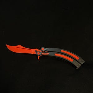 May include: A butterfly knife with a bright orange blade and accents, contrasted by a dark gray handle. The knife is open, displaying its full length and design against a black background. The blade has a unique, curved shape.