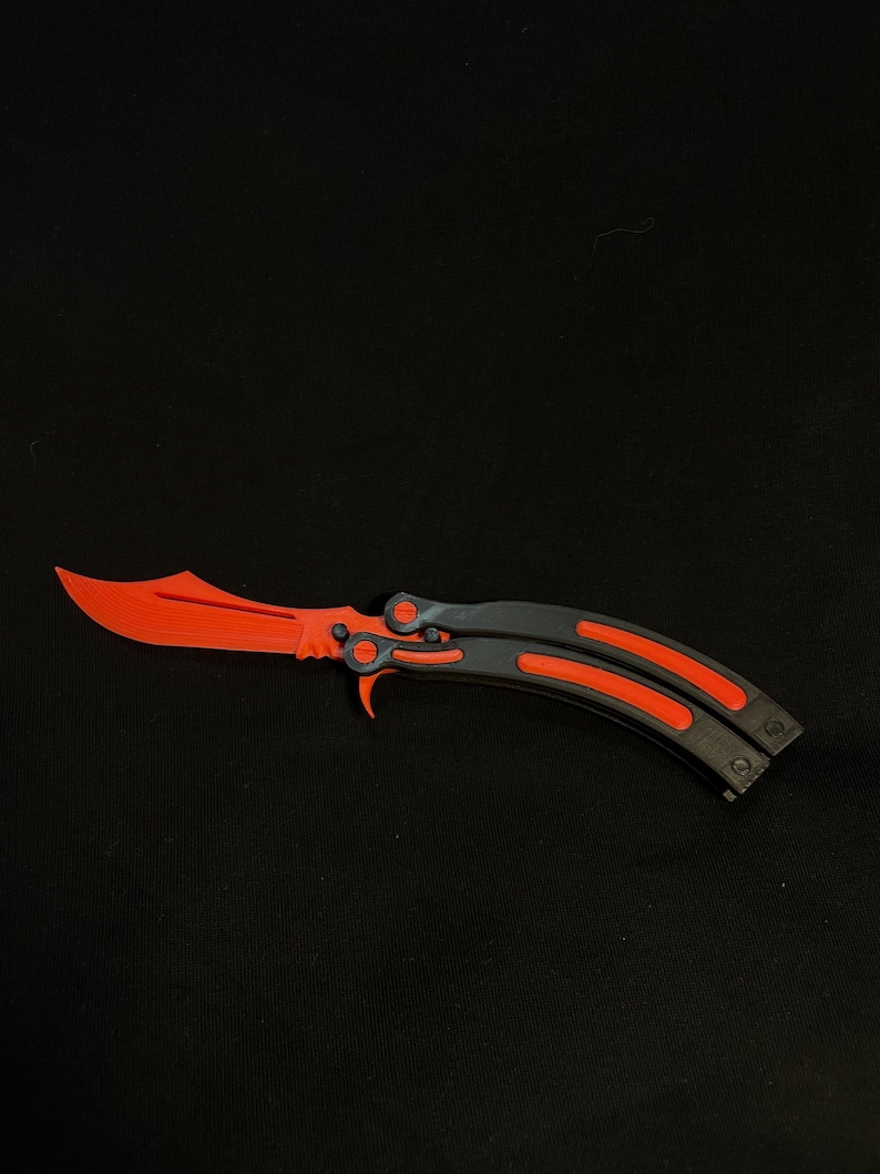 May include: A butterfly knife with a bright orange blade and accents, contrasted by a dark gray handle. The knife is open, displaying its full length and design against a black background. The blade has a unique, curved shape.