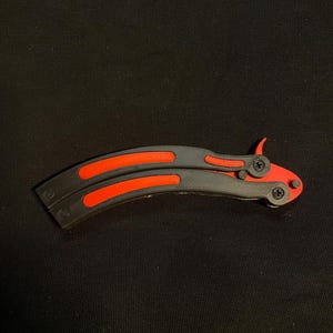 May include: A butterfly knife with a black handle and bright orange accents. The blade is curved and features a symmetrical design. The knife is closed and resting on a black surface.