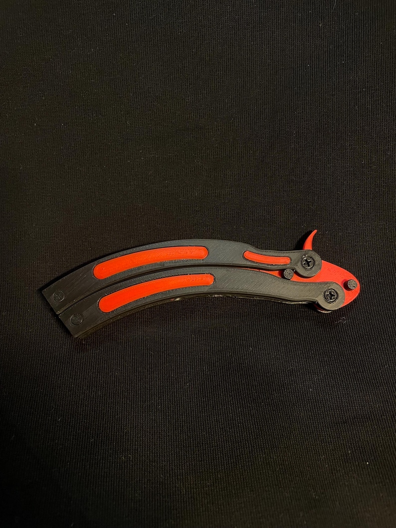 May include: A butterfly knife with a black handle and bright orange accents. The blade is curved and features a symmetrical design. The knife is closed and resting on a black surface.
