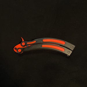 May include: A black and red butterfly knife, featuring a curved blade and handle design. The handle has a black base with red accents, and the blade is also red. The knife is lying on a black surface.