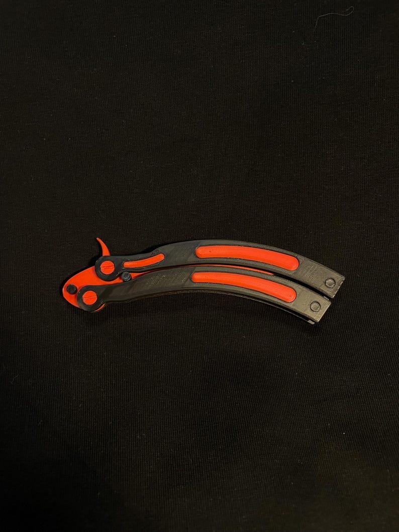 May include: A black and red butterfly knife, featuring a curved blade and handle design. The handle has a black base with red accents, and the blade is also red. The knife is lying on a black surface.
