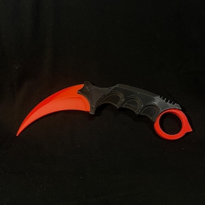 Könnte beinhalten: Ein gebogenes Karambit-Messer mit einer leuchtend roten Klinge und einem schwarzen Griff mit strukturierter Oberfläche. Der Griff verfügt über einen markanten roten Fingerring an der Basis, der eine ergonomische Form und eine spitze Klinge aufweist.