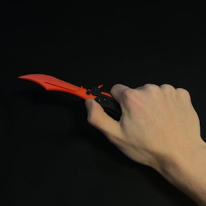 May include: A red and black butterfly knife with a curved blade. The knife is held against a black background. The blade is a bright red color, and the handle is black.