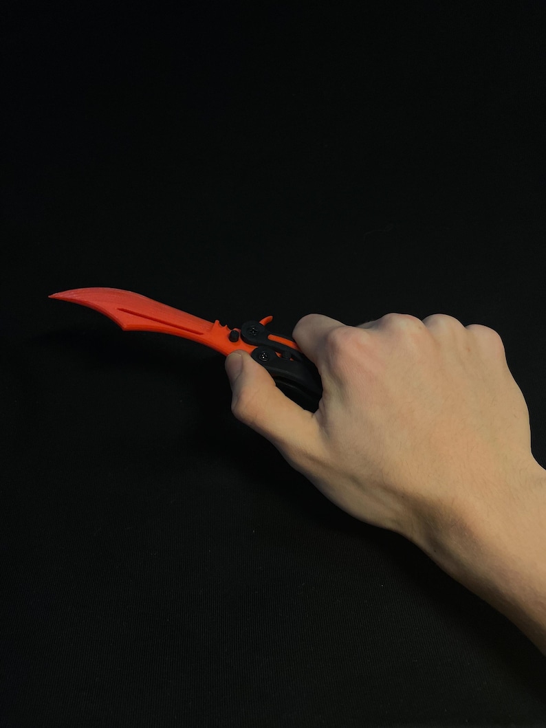 May include: A red and black butterfly knife with a curved blade. The knife is held against a black background. The blade is a bright red color, and the handle is black.