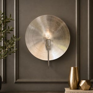 May include: A circular wall sconce with a brushed silver finish and a central vertical arm holding a single light bulb. The fixture features a large, flat, disc-shaped backplate with a metallic sheen that reflects light outward from the center.