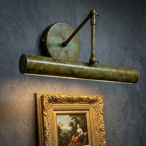 May include: A wall-mounted picture light with a cylindrical head and an adjustable arm, featuring a weathered green and bronze patina finish. Below, a gold-toned ornate frame holds a landscape painting, accompanied by two stacked books and a magnifying glass.