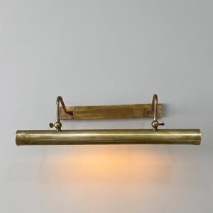 May include: A horizontal brass picture light fixture with a rectangular wall-mounting plate, two curved support arms, and a cylindrical lamp housing. The fixture features a warm, glowing light emitting from the underside of the metal tube.