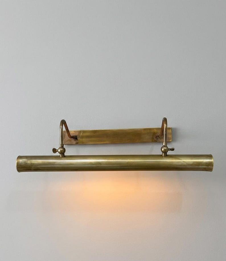 May include: A horizontal brass picture light fixture with a rectangular wall-mounting plate, two curved support arms, and a cylindrical lamp housing. The fixture features a warm, glowing light emitting from the underside of the metal tube.