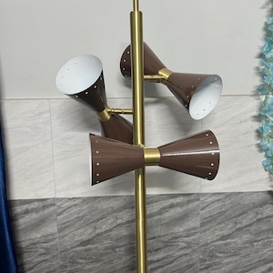 May include: A floor lamp featuring a central gold-toned metal pole with three adjustable brown cone-shaped shades. Each shade has a white interior and a series of small perforations along the rim. The lamp structure is vertical and metallic.