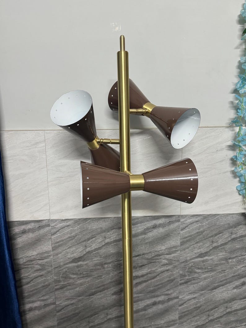 May include: A floor lamp featuring a central gold-toned metal pole with three adjustable brown cone-shaped shades. Each shade has a white interior and a series of small perforations along the rim. The lamp structure is vertical and metallic.