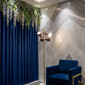 May include: A navy blue velvet armchair with a gold metal frame, a tall floor lamp with three conical brown shades on a gold pole, and deep blue floor-to-ceiling curtains adorned with cascading white and green artificial floral vines.