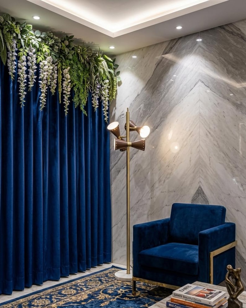 May include: A navy blue velvet armchair with a gold metal frame, a tall floor lamp with three conical brown shades on a gold pole, and deep blue floor-to-ceiling curtains adorned with cascading white and green artificial floral vines.