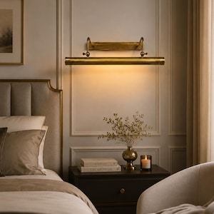 May include: A horizontal brass picture light fixture mounted on a wall, featuring a cylindrical lamp head with adjustable arms, emitting a warm downward glow. The metal finish is aged brass, and the design includes a rectangular wall plate.