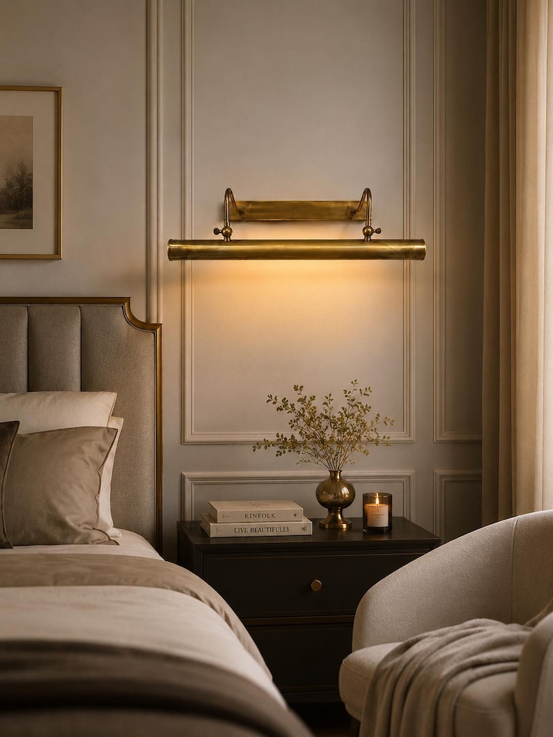 May include: A horizontal brass picture light fixture mounted on a wall, featuring a cylindrical lamp head with adjustable arms, emitting a warm downward glow. The metal finish is aged brass, and the design includes a rectangular wall plate.