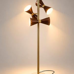 May include: A floor lamp with a tall, slender brass-colored central pole and a circular white marble base. It features four cone-shaped shades in a dark brown finish with brass accents, each emitting a warm light. A black power cord is attached.