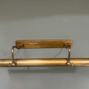 May include: A horizontal picture light fixture with a metallic brass finish. The design features a rectangular wall-mounting plate connected by two curved, adjustable arms to a long, cylindrical light housing that spans the width of the unit.