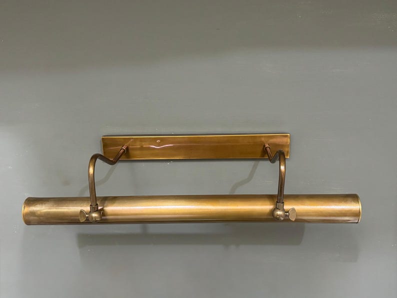 May include: A horizontal picture light fixture with a metallic brass finish. The design features a rectangular wall-mounting plate connected by two curved, adjustable arms to a long, cylindrical light housing that spans the width of the unit.