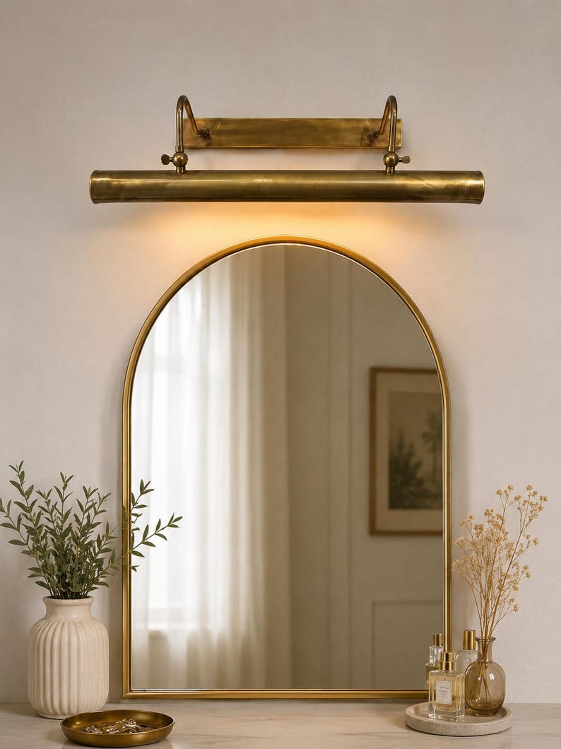May include: An arched wall mirror with a thin gold frame, positioned beneath a horizontal brass picture light fixture. The light emits a warm glow downward, illuminating the mirror surface which reflects a room interior with a framed wall art piece.