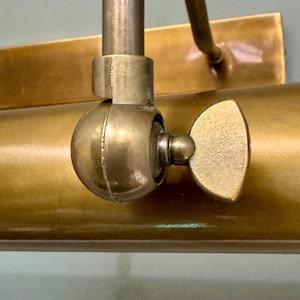 May include: A brass picture light fixture featuring a cylindrical lamp head and a rectangular wall mounting plate. The lamp head is connected to the arm by a circular swivel joint with a textured wing nut for adjusting the angle of the light.