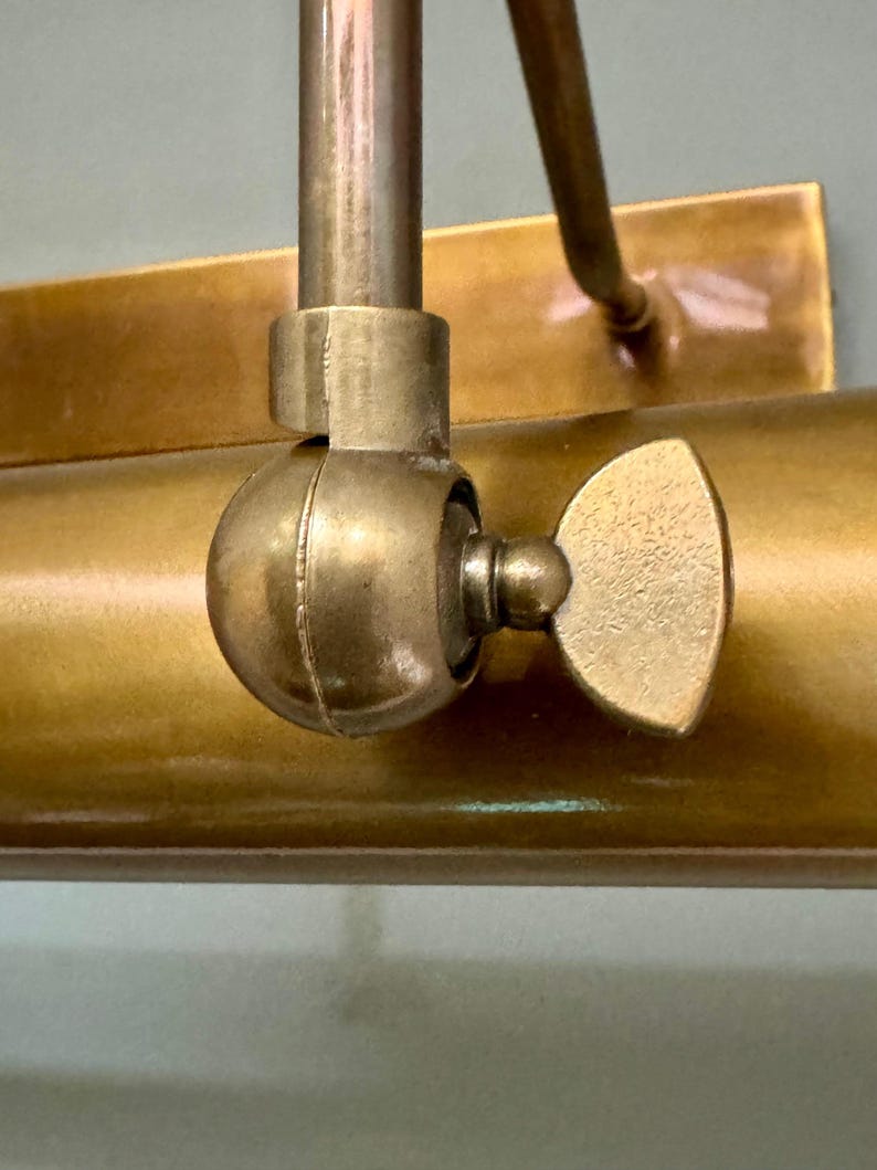 May include: A brass picture light fixture featuring a cylindrical lamp head and a rectangular wall mounting plate. The lamp head is connected to the arm by a circular swivel joint with a textured wing nut for adjusting the angle of the light.