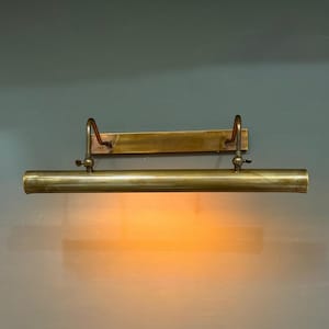 May include: A wall-mounted picture light fixture crafted from brushed brass. The horizontal cylindrical lamp head is attached to a rectangular backplate by two adjustable curved arms, emitting a warm, soft orange glow downward.