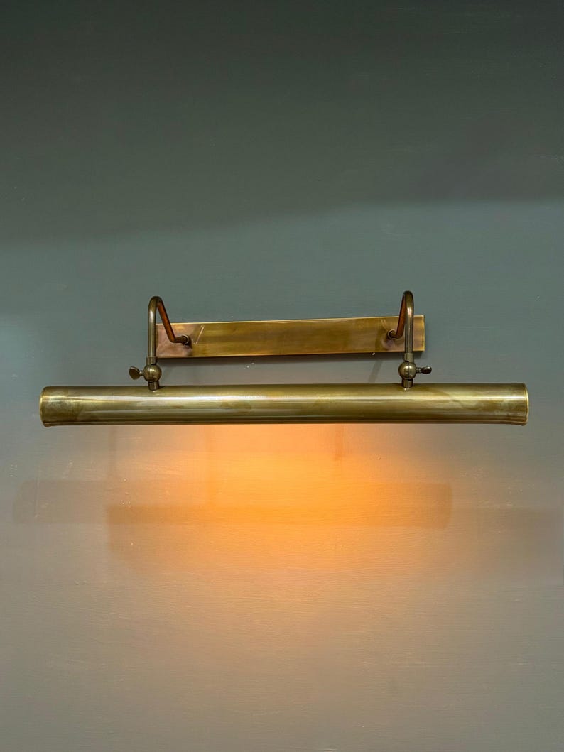 May include: A wall-mounted picture light fixture crafted from brushed brass. The horizontal cylindrical lamp head is attached to a rectangular backplate by two adjustable curved arms, emitting a warm, soft orange glow downward.