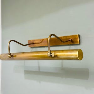 May include: A wall-mounted picture light fixture crafted from antique brass. The design features a rectangular backplate, two curved adjustable arms, and a long cylindrical light housing with visible side screws for positioning the lamp angle.