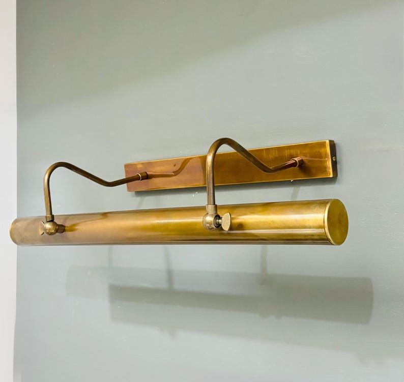May include: A wall-mounted picture light fixture crafted from antique brass. The design features a rectangular backplate, two curved adjustable arms, and a long cylindrical light housing with visible side screws for positioning the lamp angle.