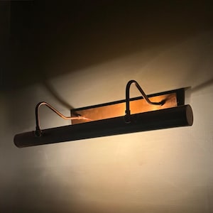 May include: A horizontal wall-mounted picture light fixture with a metallic copper finish. The lamp features a long cylindrical shade supported by two curved arms attached to a rectangular backplate, emitting a warm glow against the wall.