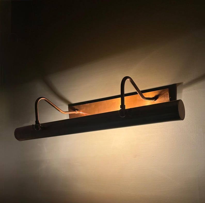 May include: A horizontal wall-mounted picture light fixture with a metallic copper finish. The lamp features a long cylindrical shade supported by two curved arms attached to a rectangular backplate, emitting a warm glow against the wall.