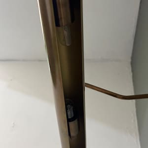 May include: A long, cylindrical, metallic gold-toned light fixture with a hollow interior channel. Two small, clear glass light bulbs are mounted inside the vertical housing. A thin, angled metal arm extends from the side of the main structure.