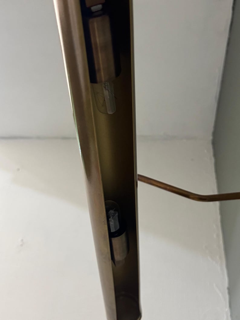 May include: A long, cylindrical, metallic gold-toned light fixture with a hollow interior channel. Two small, clear glass light bulbs are mounted inside the vertical housing. A thin, angled metal arm extends from the side of the main structure.