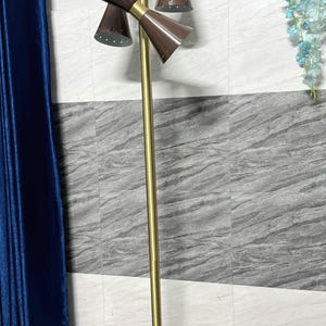 May include: A floor lamp featuring a tall, slender gold-toned metal pole mounted on a circular white base. The lamp has three brown, cone-shaped metal shades with small perforated holes along the edges, arranged at different heights and angles.