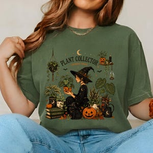 May include: An olive green t-shirt featuring a graphic print of a witch surrounded by various potted plants, a black cat, and pumpkins. The text reads "PLANT COLLECTOR" and "GROWING MAGIC" in a dark font, accompanied by moon and star illustrations.