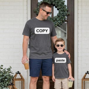 May include: Two matching gray t-shirts, one with "COPY" and the other with "paste" in white text. The shirts are worn by a man and a child, both wearing sunglasses. The man is wearing navy shorts, and the child is wearing khaki shorts.