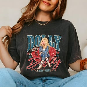 May include: A charcoal gray short-sleeve t-shirt featuring a graphic of a blonde woman in a denim outfit and cowboy boots. The design includes a large red heart, blue stars, and the text DOLLY DOLLY 1946-2026 in a bold, distressed font style.